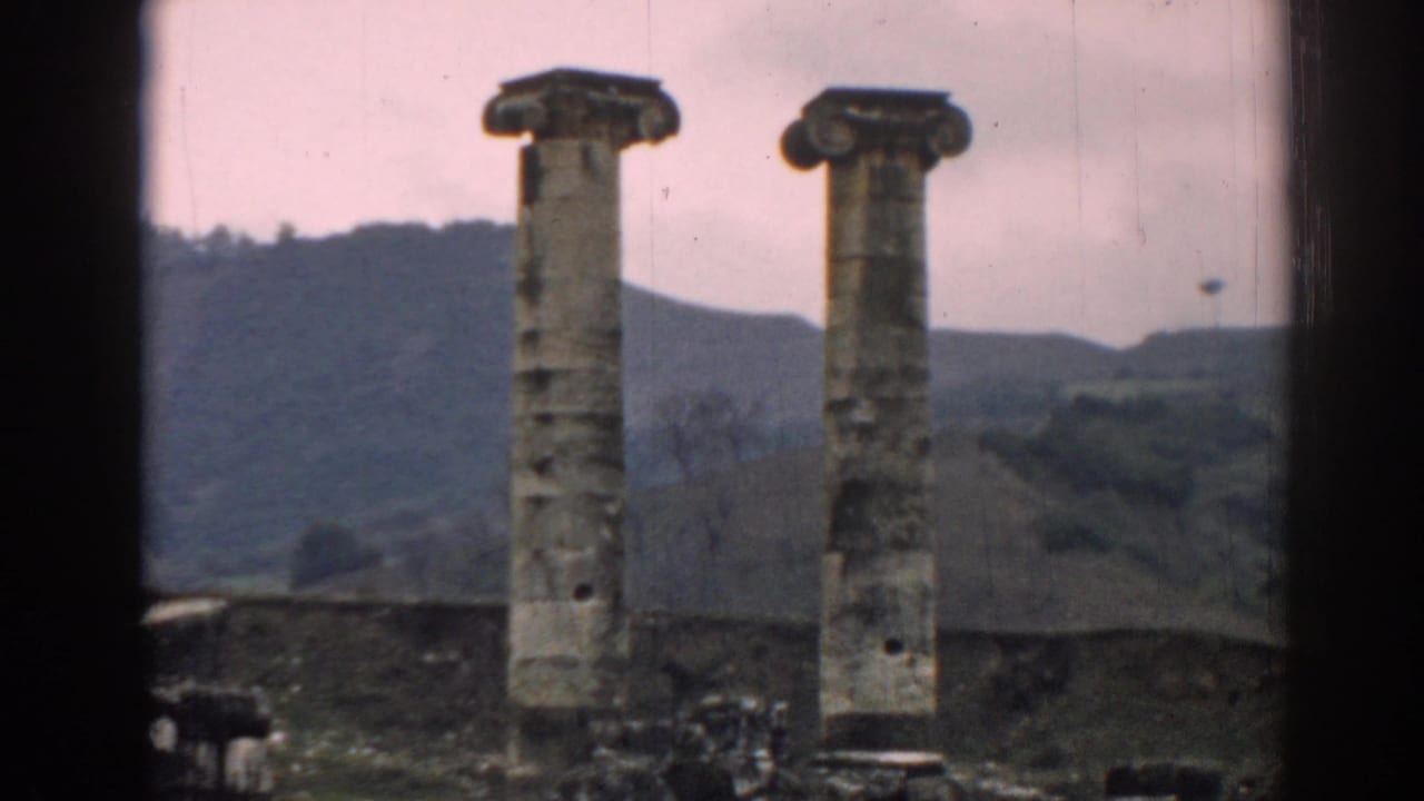 Two tall stone columns stand among ancient ruins with hills behind them.