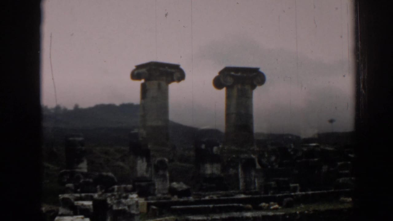Darker 1962 home-movie frame of Ephesus ruins, showing two columns, broken masonry, and a low hillside under muted light.