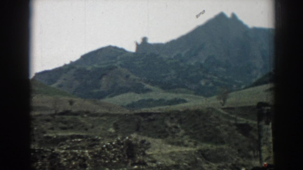 Ephesus-area hillside view on 8mm film, showing a jagged ridge, muted green slopes, cloudy sky, and foreground stonework.