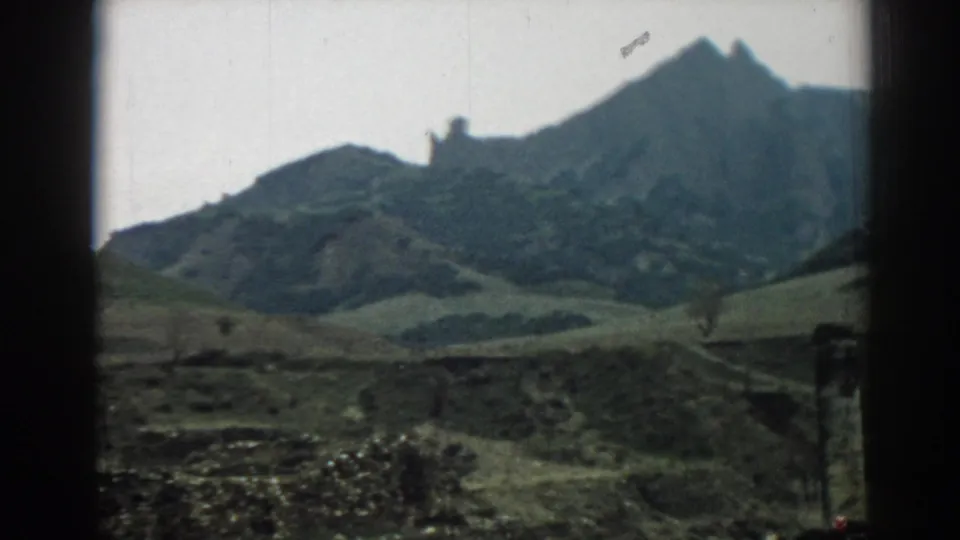 Ephesus-area hillside view on 8mm film, showing a jagged ridge, muted green slopes, cloudy sky, and foreground stonework.
