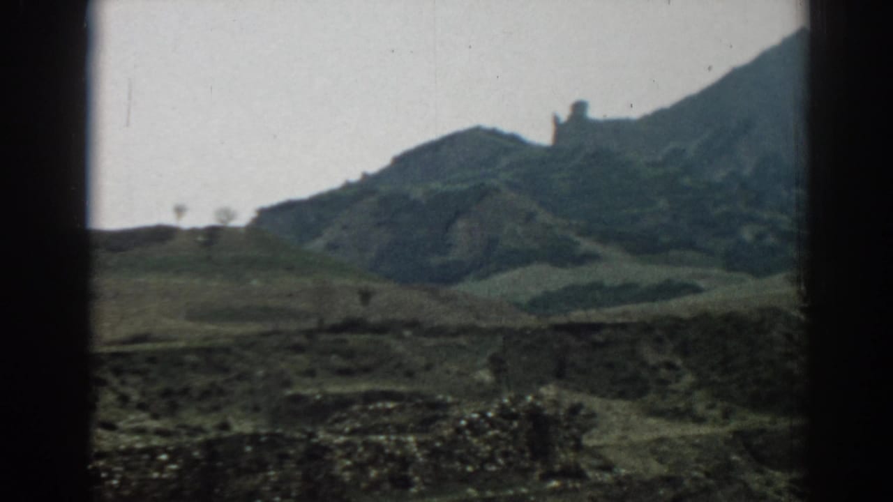 Muted 8mm travel-film view of rolling hills near Ephesus, with low stonework, cloudy light, and a partially cropped ridge.