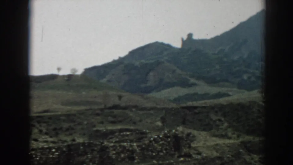 Muted 8mm travel-film view of rolling hills near Ephesus, with low stonework, cloudy light, and a partially cropped ridge.