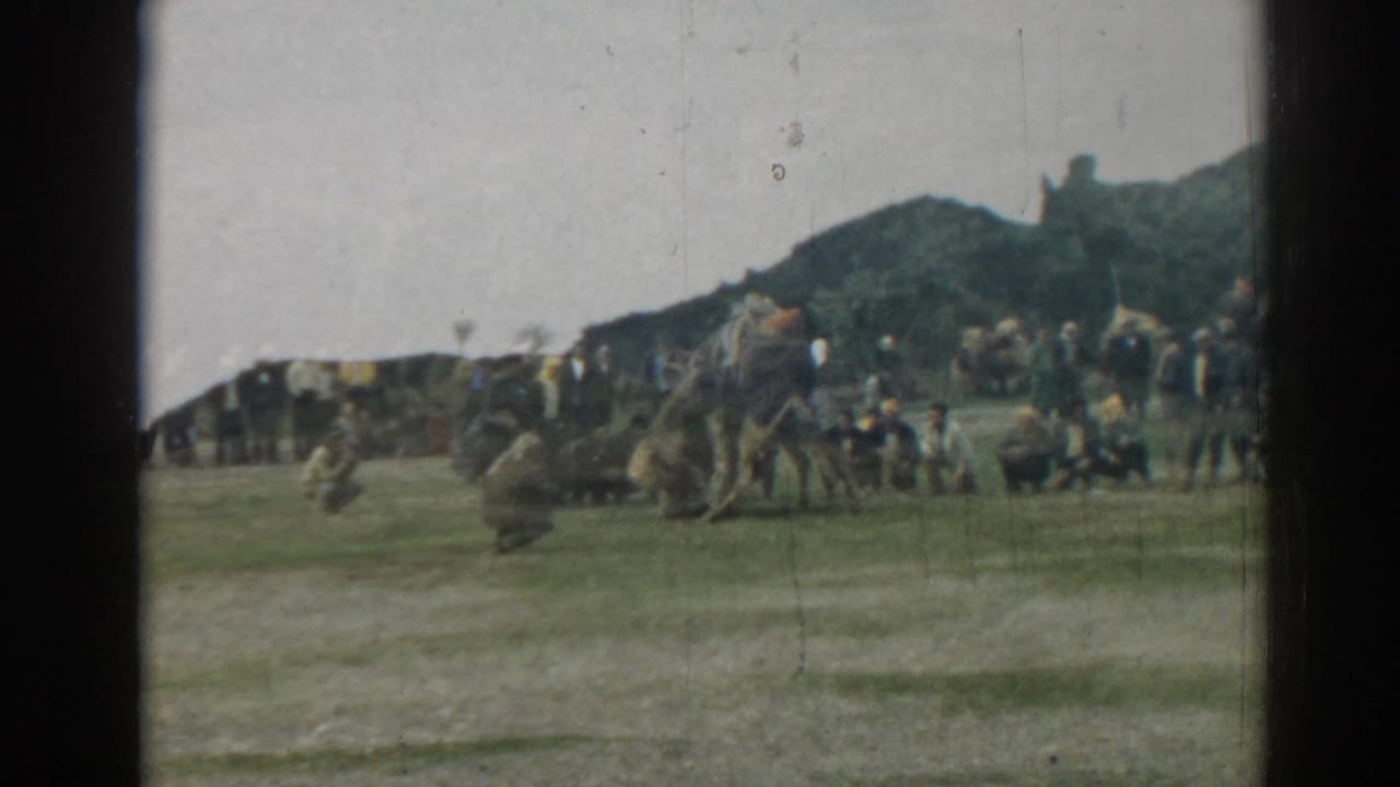 1962 Ephesus travel footage frame showing low ruins, green hills, cloudy sky, and small distant visitors without identifiable faces.
