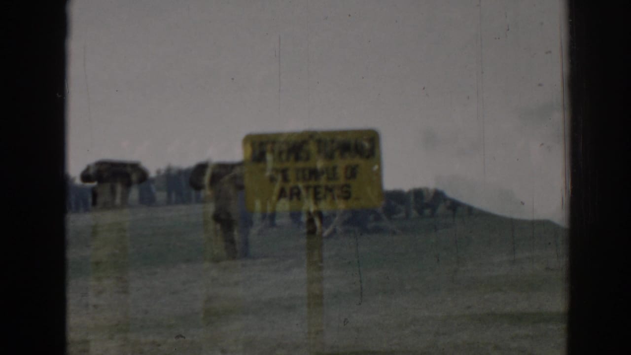 Yellow Temple of Artemis sign beside a road with parked cars and distant hills.
