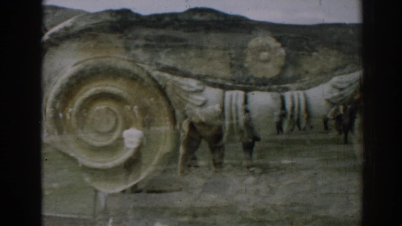 Two men and a tan bear near large carved stone ruins under a cloudy sky.
