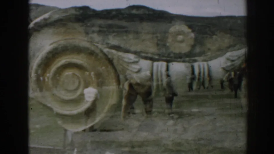 1962 Ephesus home-movie frame with two men walking a tan bear past spiral-patterned stonework and distant hills.