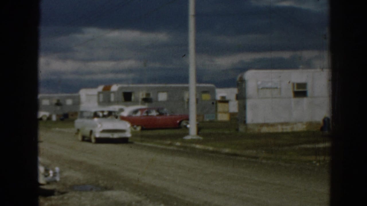 White station wagon approaches a dirt road turn near trailers and parked cars.