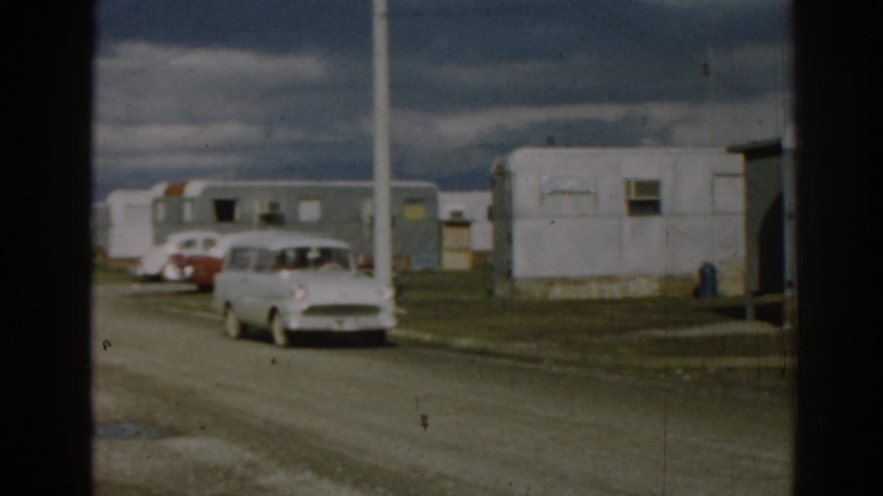 1962 Ephesus home-movie frame with the station wagon entering the park road beside trailers, parked cars, and tall poles.