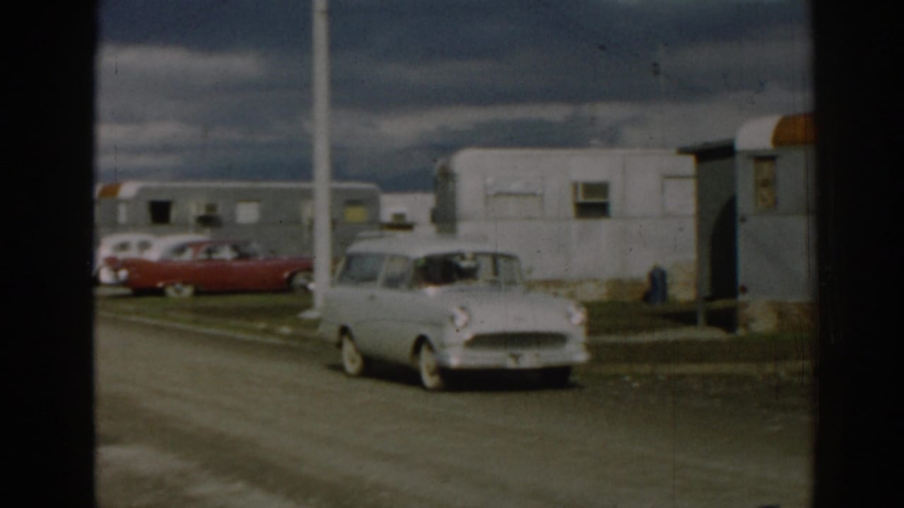 The white station wagon passes nearer to the camera in 1962 Ephesus, with a trailer row, red sedan, and cloudy sky behind it.