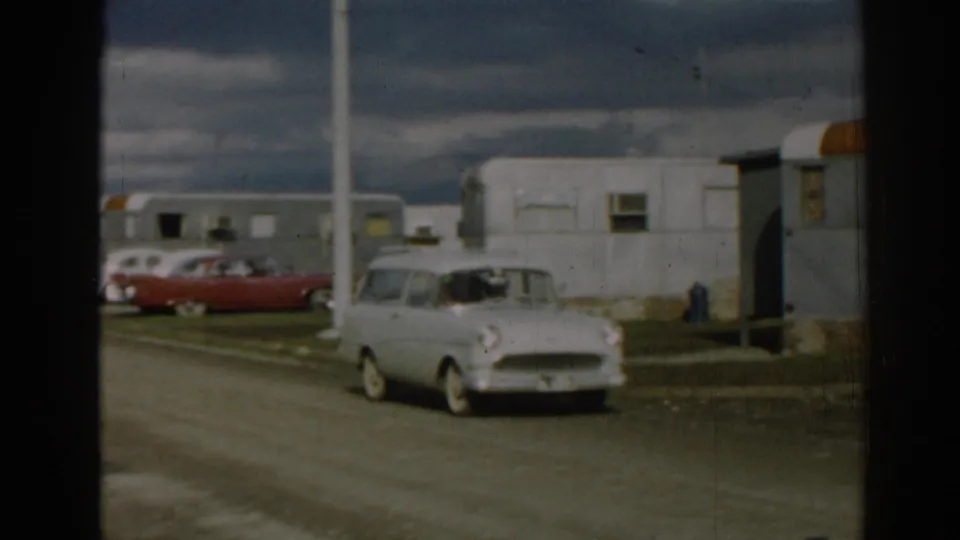 The white station wagon passes nearer to the camera in 1962 Ephesus, with a trailer row, red sedan, and cloudy sky behind it.