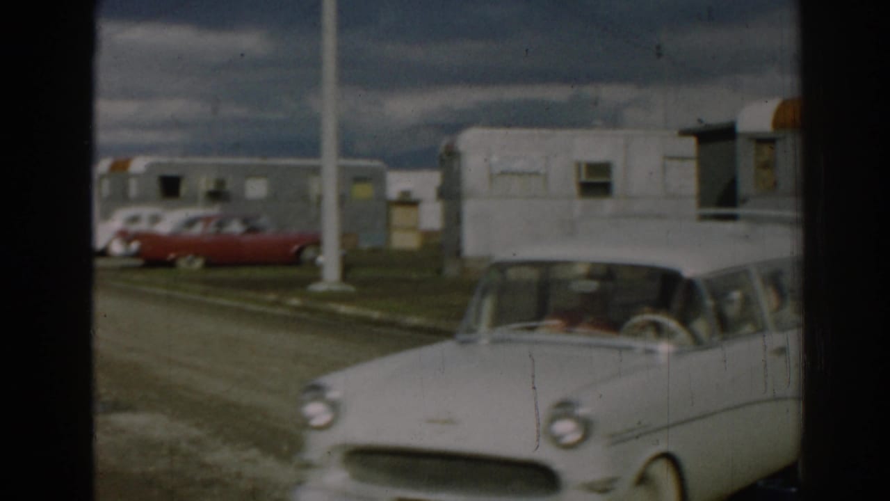 Close 1962 Ephesus frame of the station wagon passing the camera, with occupants visible and trailers and parked cars in the background.