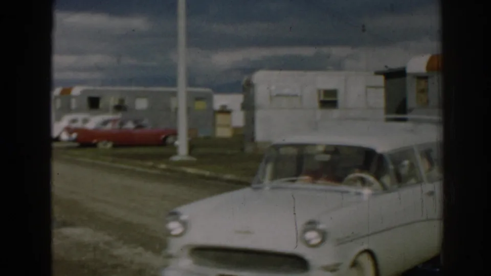 Close 1962 Ephesus frame of the station wagon passing the camera, with occupants visible and trailers and parked cars in the background.