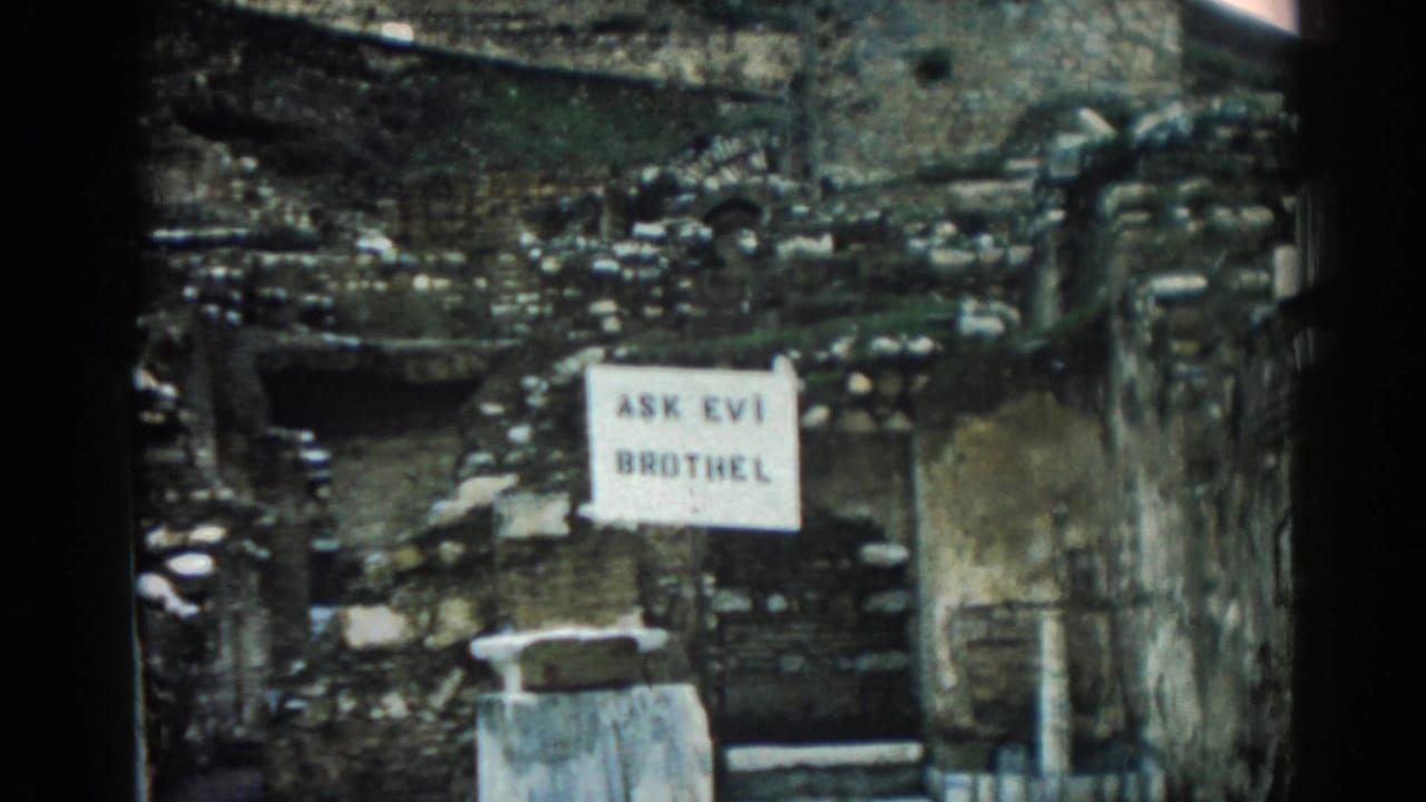 1962 Ephesus frame with a white posted sign against rough masonry and debris, filmed in faded 8mm color.