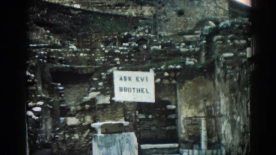 1962 Ephesus frame with a white posted sign against rough masonry and debris, filmed in faded 8mm color.