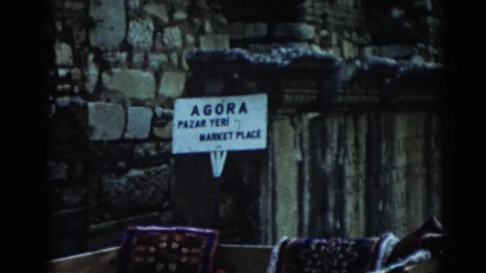 1962 Ephesus home-movie frame showing an Agora marketplace sign against ancient stonework and column fragments.