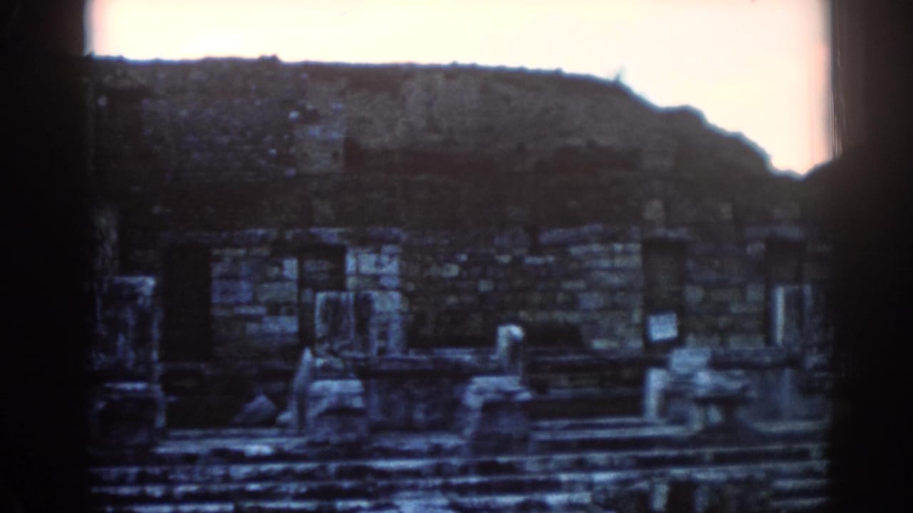 1962 8mm view of weathered stone ruins at Ephesus, with low walls, steps, and a dark hillside beyond.