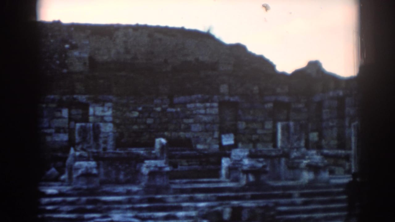 Visitor-shot 8mm frame of Ephesus ruins, showing crumbling stone walls, short columns, steps, and hillside background.
