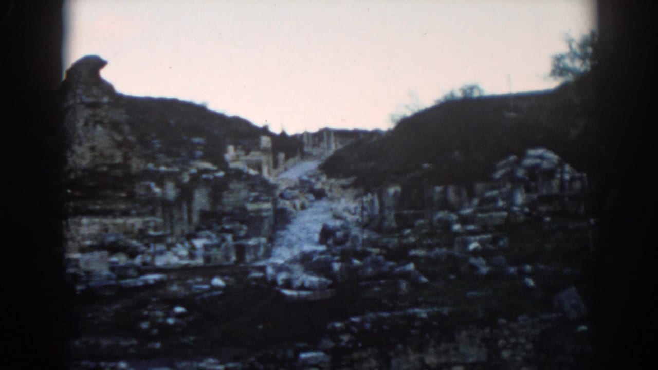1962 8mm view down a path among Ephesus ruins, with broken masonry, hillside edges, and a small visitor figure visible.