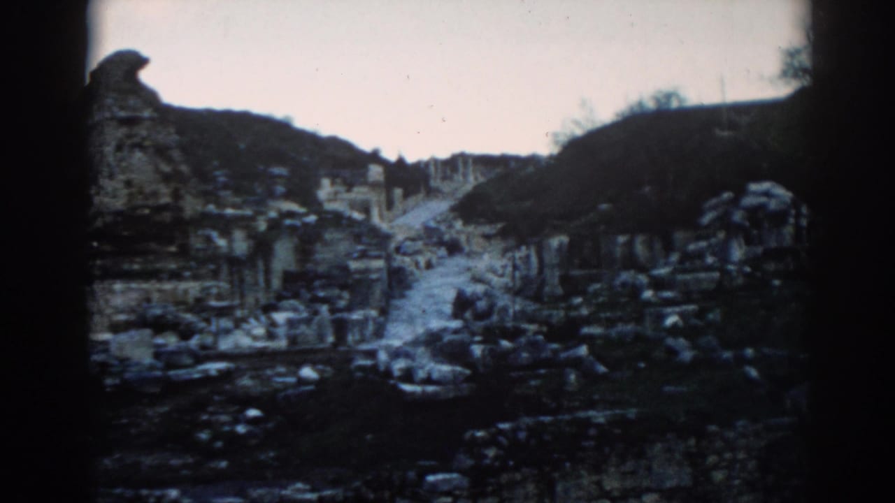 Wide 8mm frame of Ephesus ruins in 1962, with a central path, broken stone structures, and dark hills framing the site.