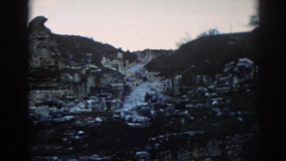Wide 8mm frame of Ephesus ruins in 1962, with a central path, broken stone structures, and dark hills framing the site.