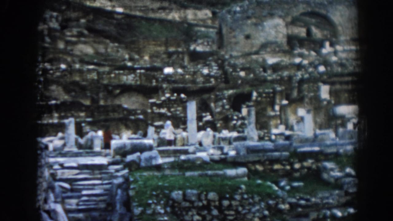 1962 home-movie frame of Ephesus showing columns, layered stone walls, steps, green grass, and a distant visitor in red.