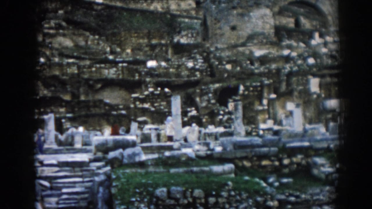 Soft 8mm frame of Ephesus ruins, with broken columns, stone steps, arched masonry, grass, and a small red-clad visitor for scale.