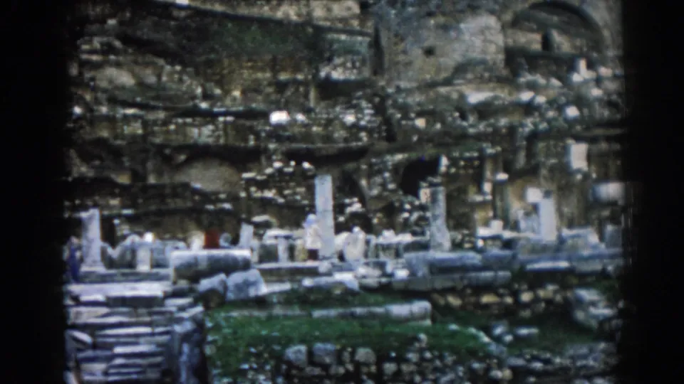 Soft 8mm frame of Ephesus ruins, with broken columns, stone steps, arched masonry, grass, and a small red-clad visitor for scale.