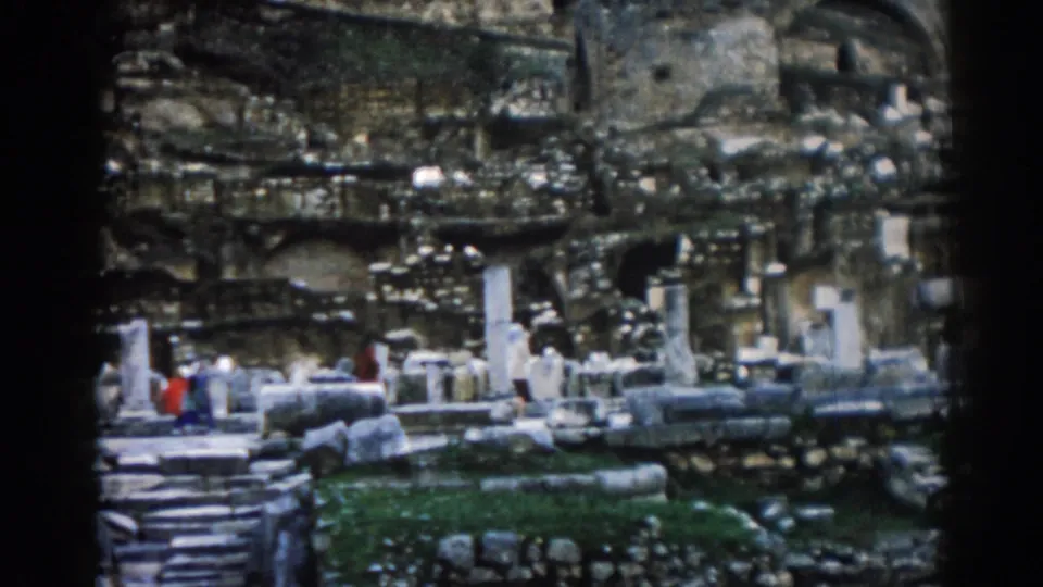 1962 Ephesus home-movie frame showing visitors moving through ancient ruins, with columns, stone steps, and weathered walls around them.