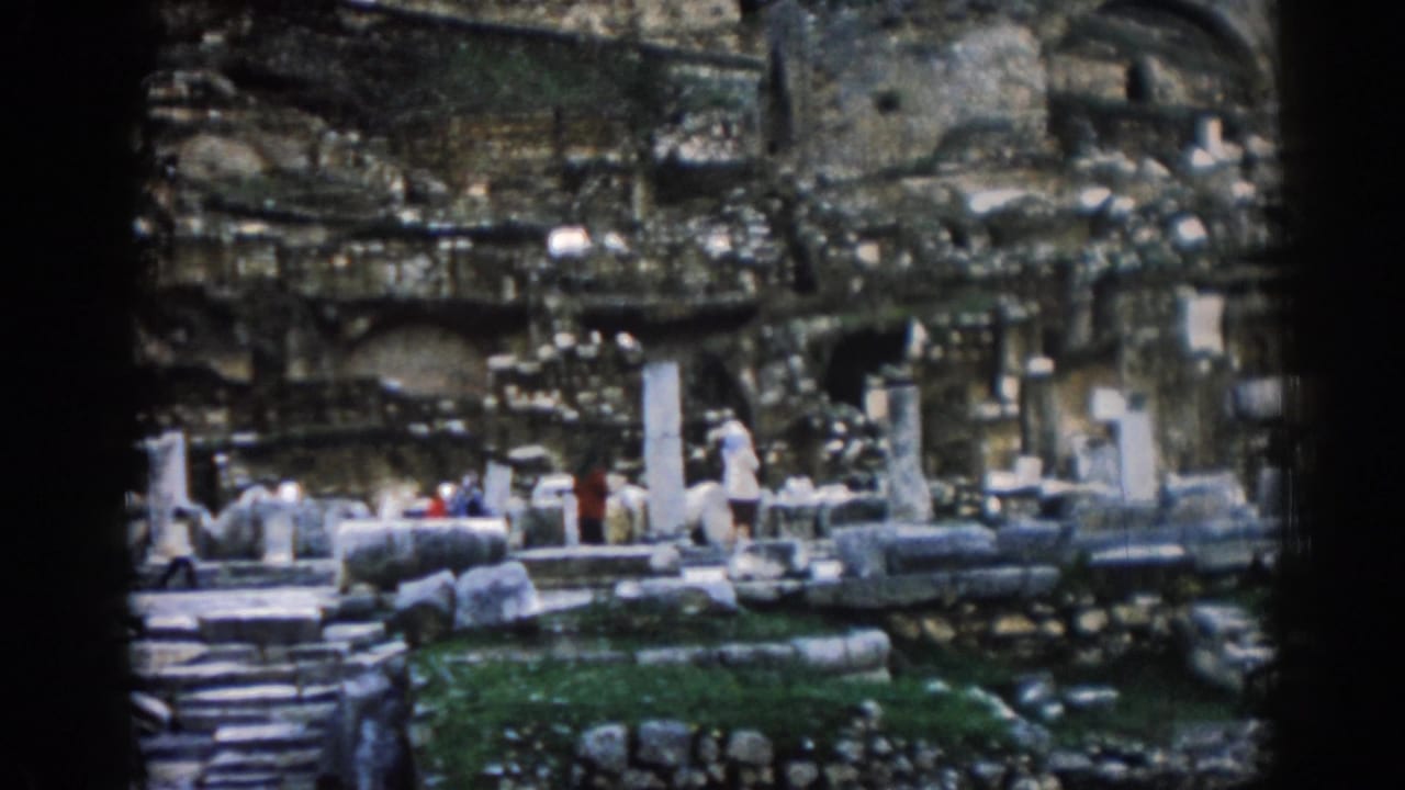 Visitors stand and walk through stone ruins beneath a weathered rock and masonry backdrop.