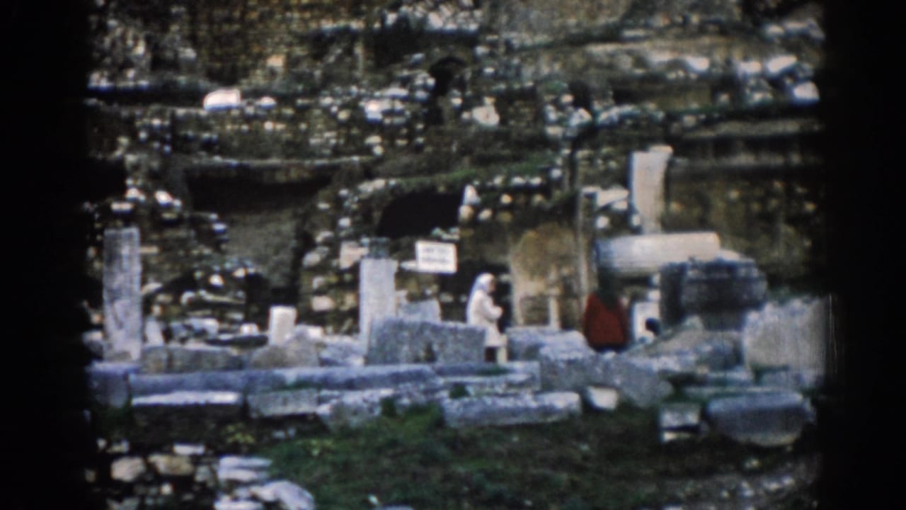 1962 Ephesus frame showing people near a small site sign, broken columns, stone blocks, grass, and shadowed openings in the ancient ruins.