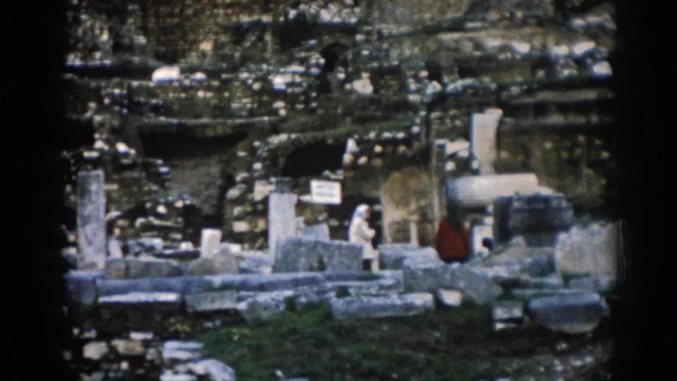 1962 Ephesus frame showing people near a small site sign, broken columns, stone blocks, grass, and shadowed openings in the ancient ruins.