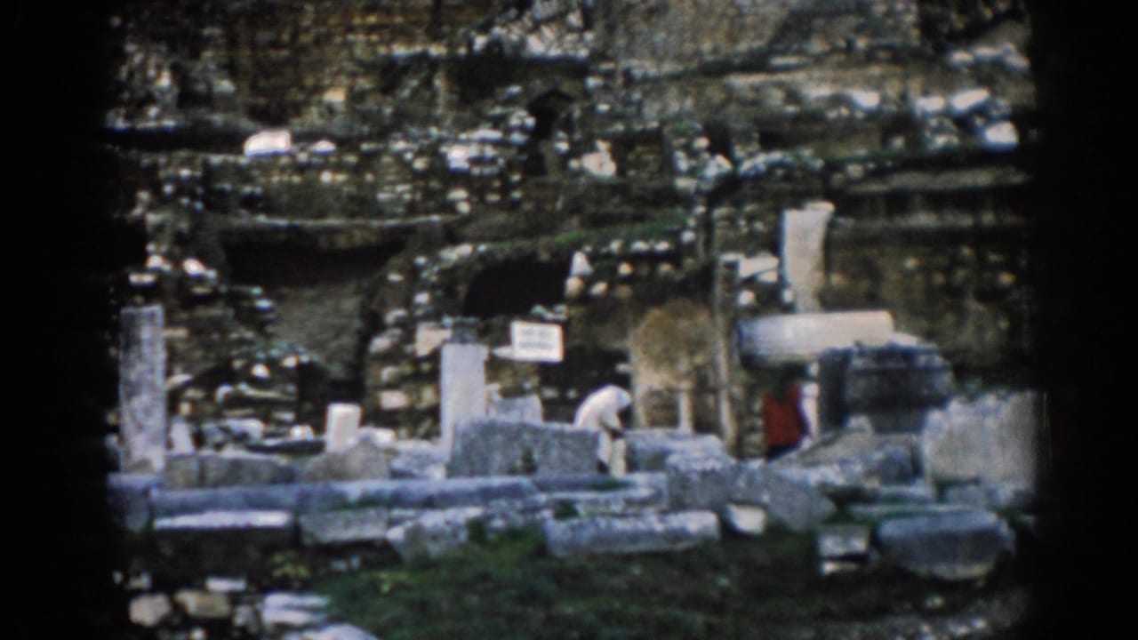 1962 Ephesus home-movie frame with visitors among broken stone blocks, upright columns, a small sign, grass, and weathered ruin walls.