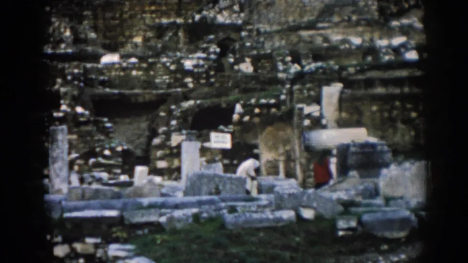 1962 Ephesus home-movie frame with visitors among broken stone blocks, upright columns, a small sign, grass, and weathered ruin walls.