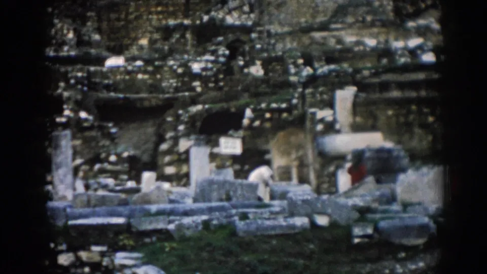 1962 8mm view of Ephesus ruins with weathered stone blocks, upright columns, mossy patches, and small distant visitors.