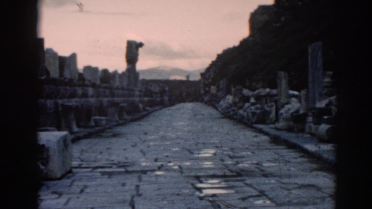Faded 8mm frame of an Ephesus cobblestone walkway bordered by columns, stone piles, hillside terrain, and a distant upright form.