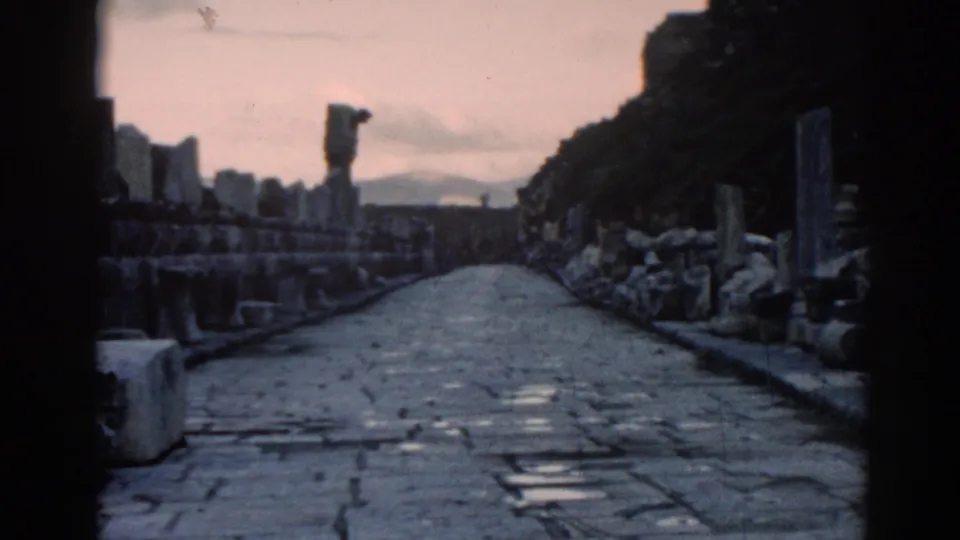 Faded 8mm frame of an Ephesus cobblestone walkway bordered by columns, stone piles, hillside terrain, and a distant upright form.