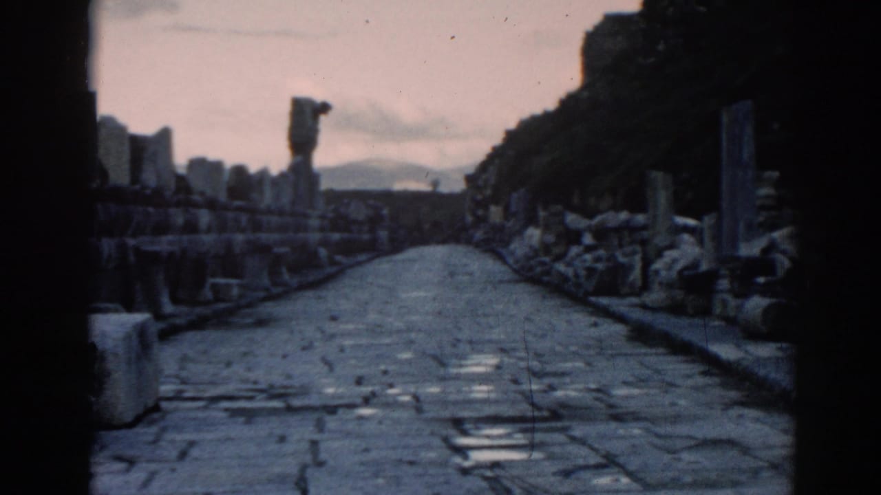 Visitor-shot 8mm frame of the Ephesus stone roadway, with columns, rubble, and dark ruin edges in view.