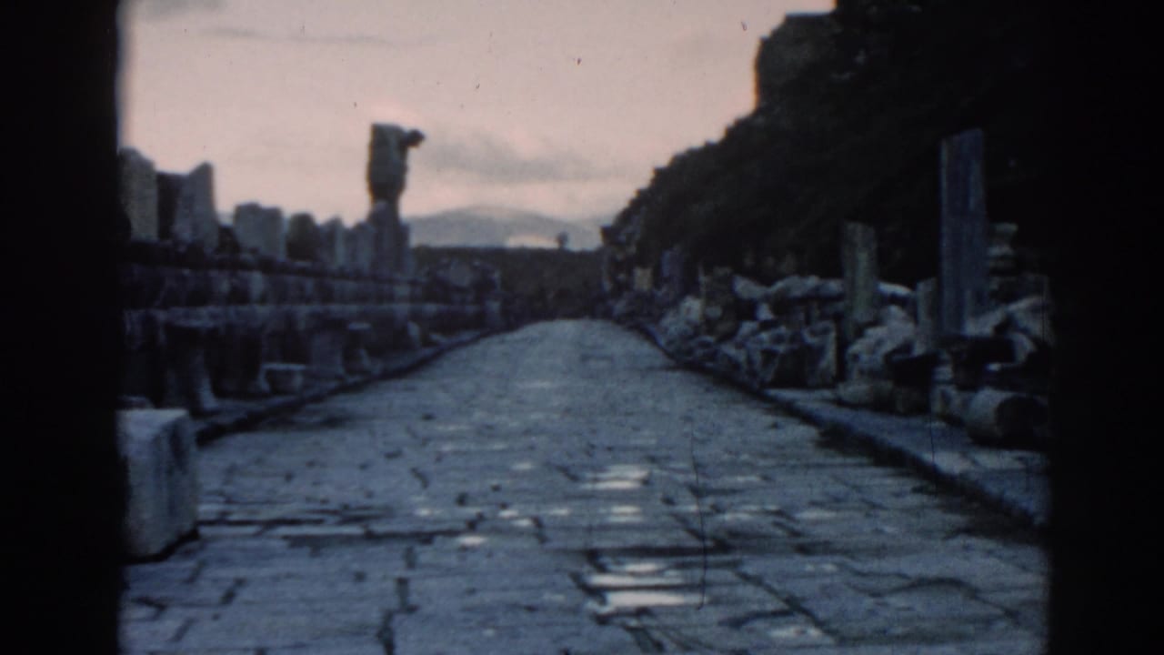 1962 Ephesus travel frame showing a stone road through ruins, with masonry fragments and a distant hazy landscape.