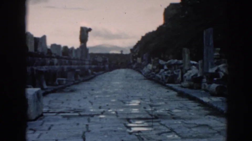 1962 Ephesus travel frame showing a stone road through ruins, with masonry fragments and a distant hazy landscape.