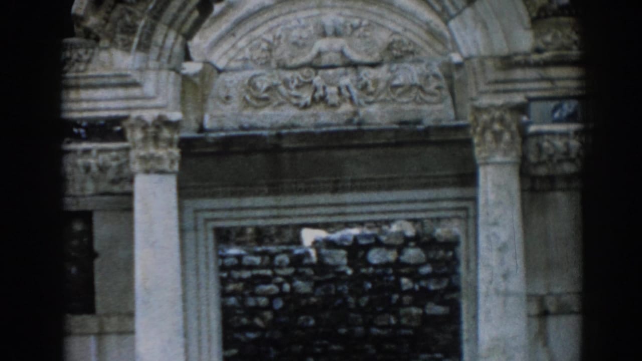 Stone archway with carved pediment, columns, and a dark rubble wall behind it.