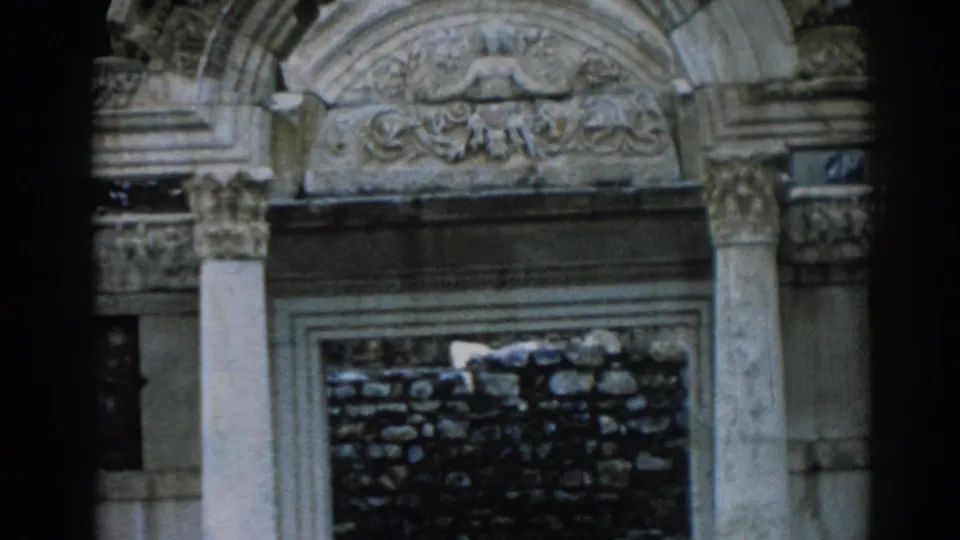 1962 Ephesus home-movie frame showing a carved stone pediment, pale columns, and a dark stone wall behind an ancient archway.