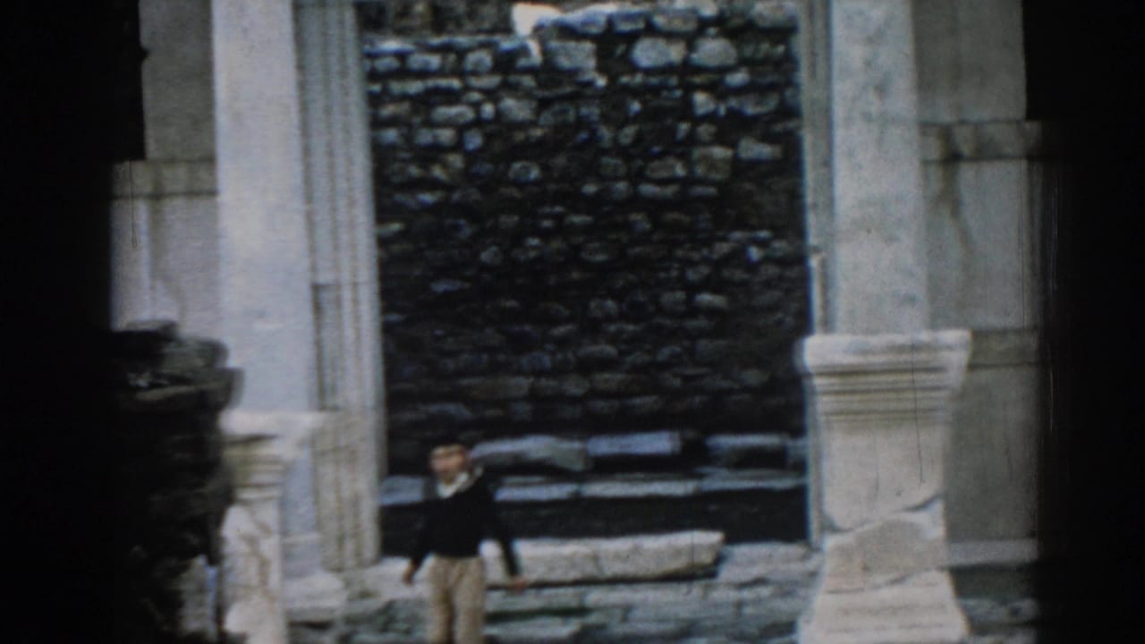 1962 Ephesus home-movie frame showing a man walking below pale columns and a dark stone wall in the ruins.