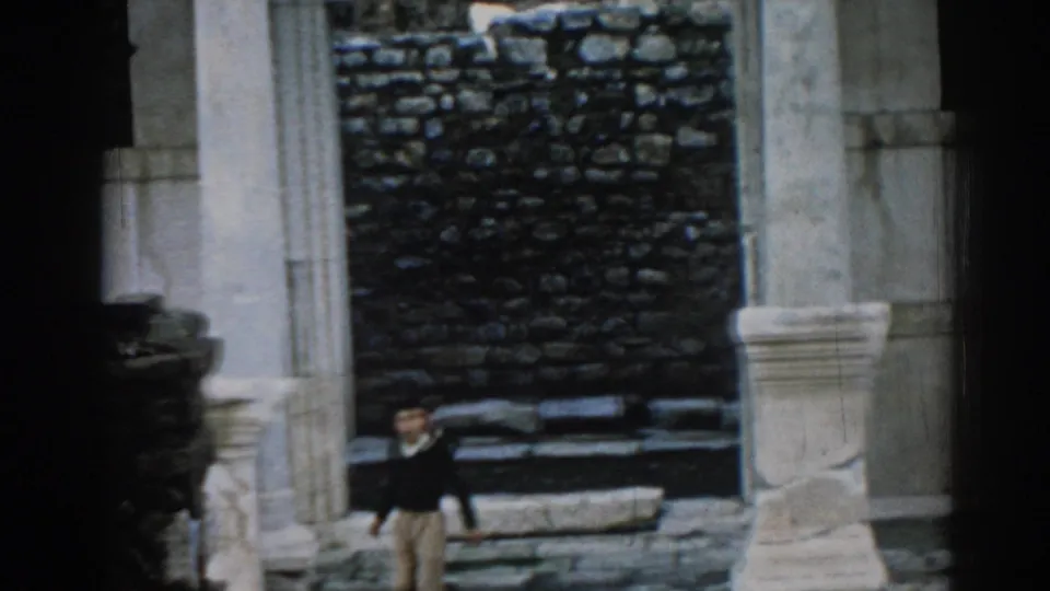 1962 Ephesus home-movie frame showing a man walking below pale columns and a dark stone wall in the ruins.