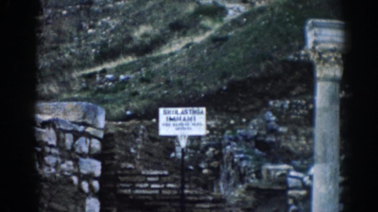 Visitor-shot 1962 Ephesus frame showing a site sign among stone ruins, columns, steps, and a grassy hillside.