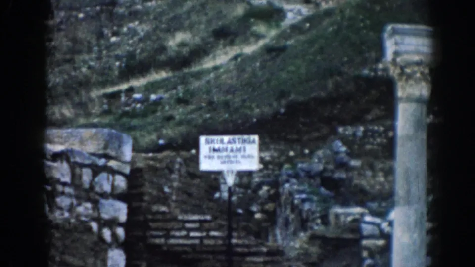 Visitor-shot 1962 Ephesus frame showing a site sign among stone ruins, columns, steps, and a grassy hillside.