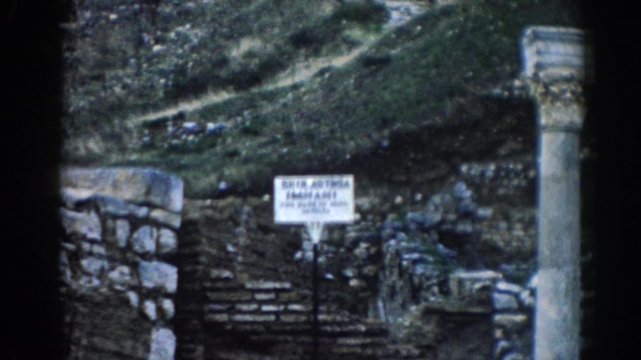 1962 8mm frame at Ephesus showing weathered ruins, a hillside path, a white site sign, and a tall stone column.