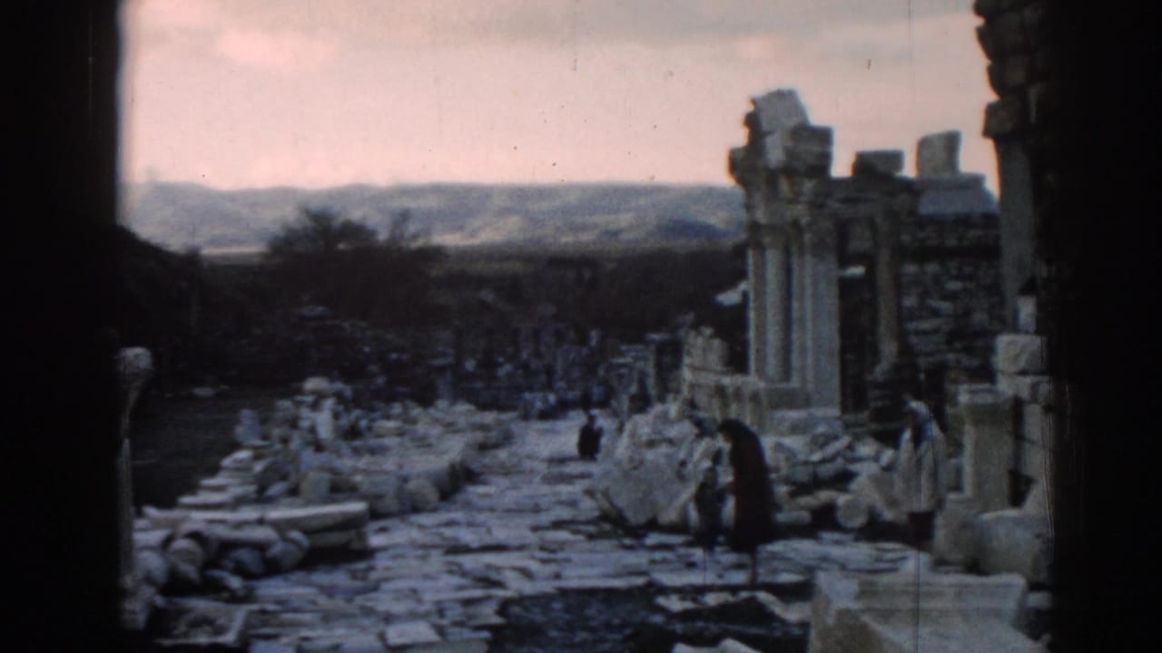 Wide 1962 Ephesus view with column remains, rubble-lined path, distant hills, and visitors moving through the ruins.