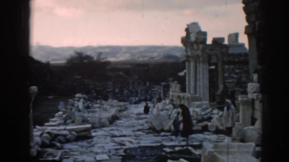Wide 1962 Ephesus view with column remains, rubble-lined path, distant hills, and visitors moving through the ruins.