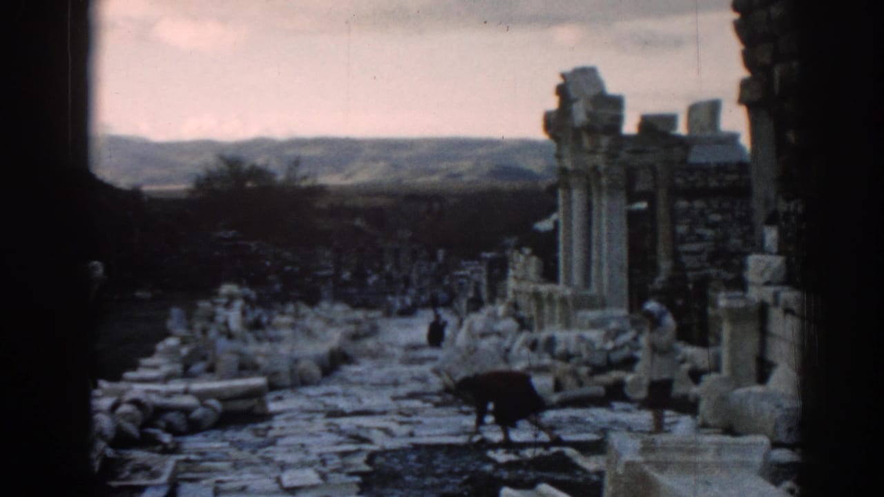 1962 Ephesus travel footage frame showing visitors walking through a rubble-lined path beside columns and ruined stone walls.