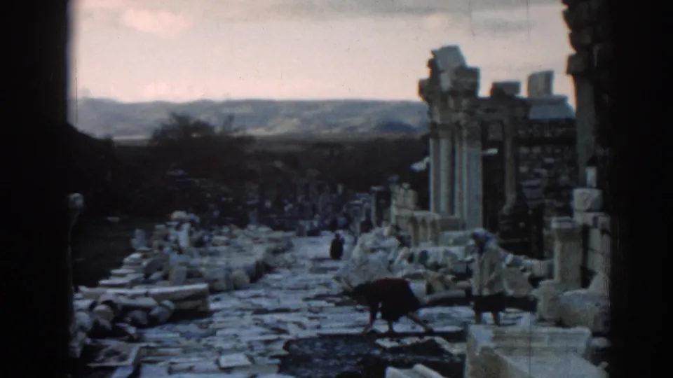1962 Ephesus travel footage shows visitors among stone columns, rubble, and a path framed by dark foreground edges.